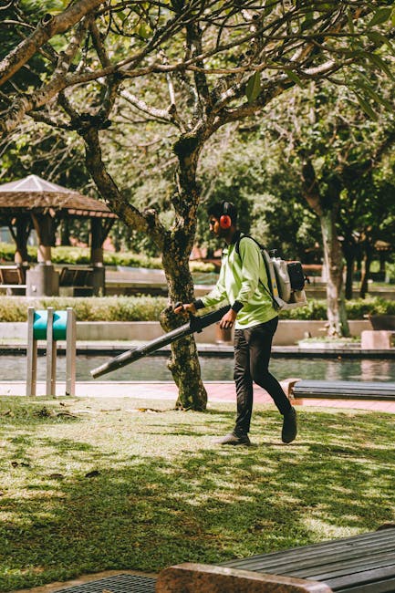 A person wearing a light green jacket, black trousers, and red ear protection is using a leaf blower to tidy up fallen leaves and debris in a well-maintained garden. The garden features a large, matur