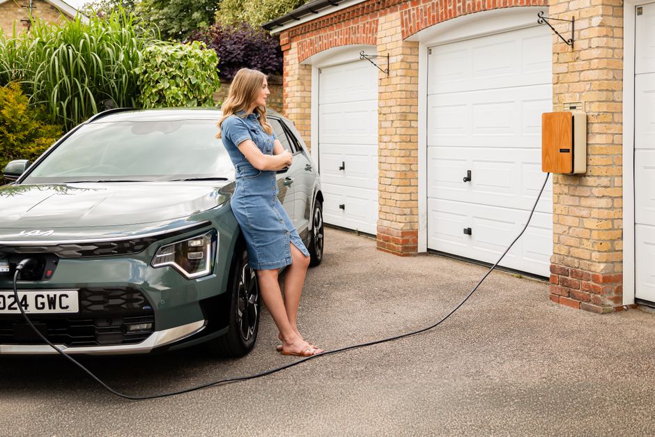 A young woman with shoulder-length blonde hair, dressed in a casual blue denim dress and sandals, stands beside a modern grey electric car parked in a driveway. She is holding a smartphone in her righ